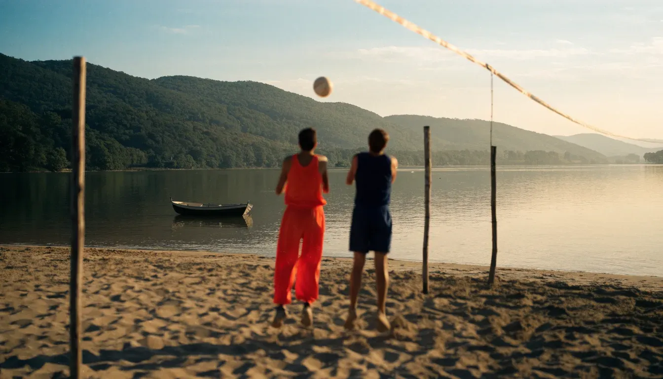 Ein Volleyball liegt im Sand eines Spielfelds am Seeufer, im Hintergrund gleitet ein Boot über das Wasser.