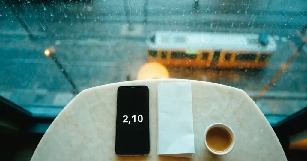 Ein Café-Tisch am Fenster trägt ein Smartphone mit dunklem Display, an der Scheibe laufen Regentropfen herunter und draussen steht eine Tram.