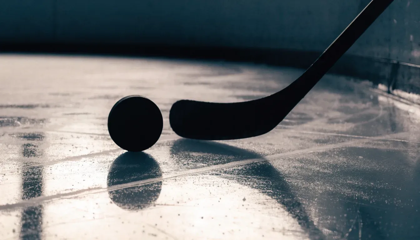 Ein Eishockeypuck liegt auf der Eisfläche neben dem Blatt eines Schlägers, die Bande dahinter ist im Dunkeln nur schwach zu erkennen.