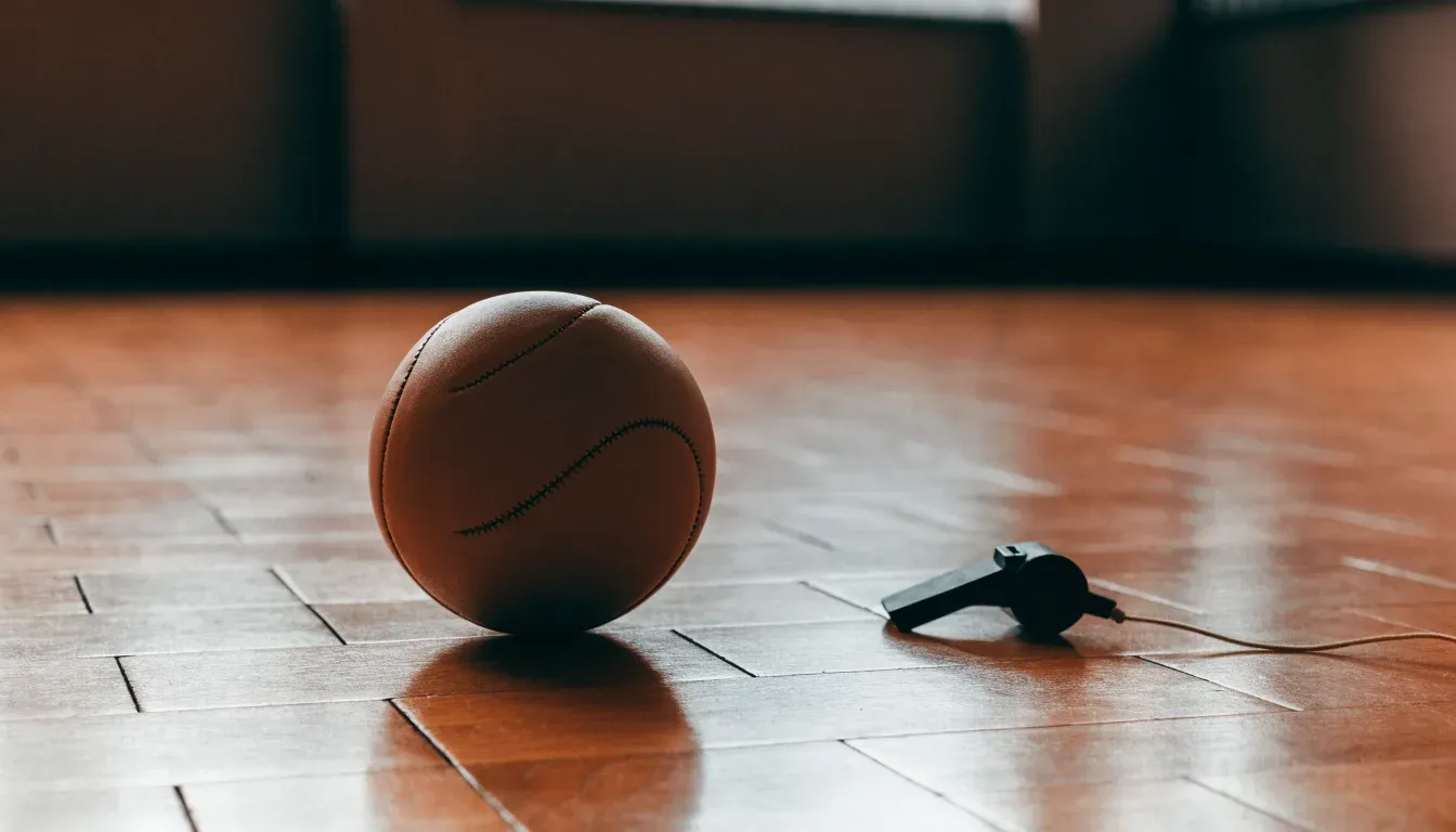 Ein kleiner, schwerer Ball liegt auf glattem Hallenparkett neben einer Trillerpfeife an einer Schnur.