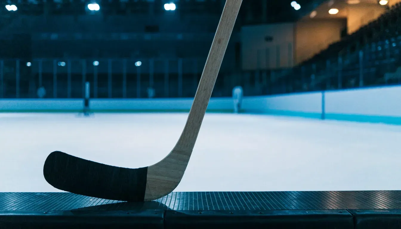 Ein Eishockeyschläger lehnt an der Bande, in der Ferne liegt die hell beleuchtete Eisfläche einer Halle unter Flutlicht.