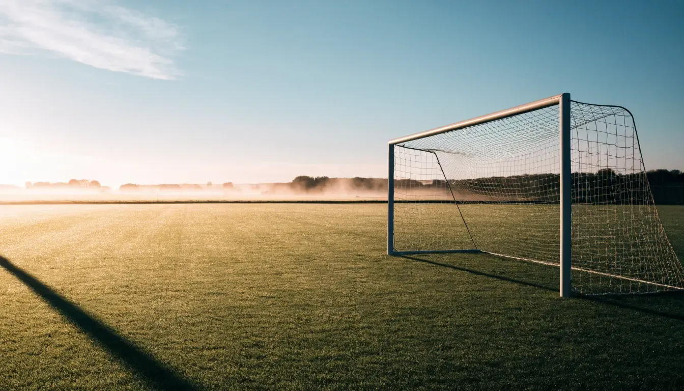 Ein Fussballtor mit Netz in der Totalen auf einem leeren Platz, Tau glänzt im Gras und die Morgensonne steht tief.