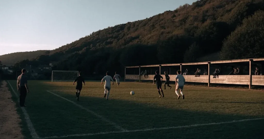 Ein kleiner Fussballplatz mit einfacher Holztribüne und Rasenstreifen am Rand einer Ortschaft, im Hintergrund ein bewaldeter Hügel im Abendlicht.