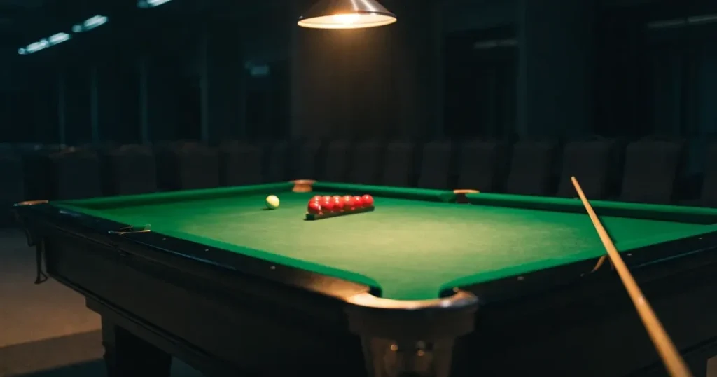 Ein Snookertisch mit grünem Tuch und einem Dreieck aus roten Kugeln unter einer tief hängenden Lampe in einer leeren Halle.