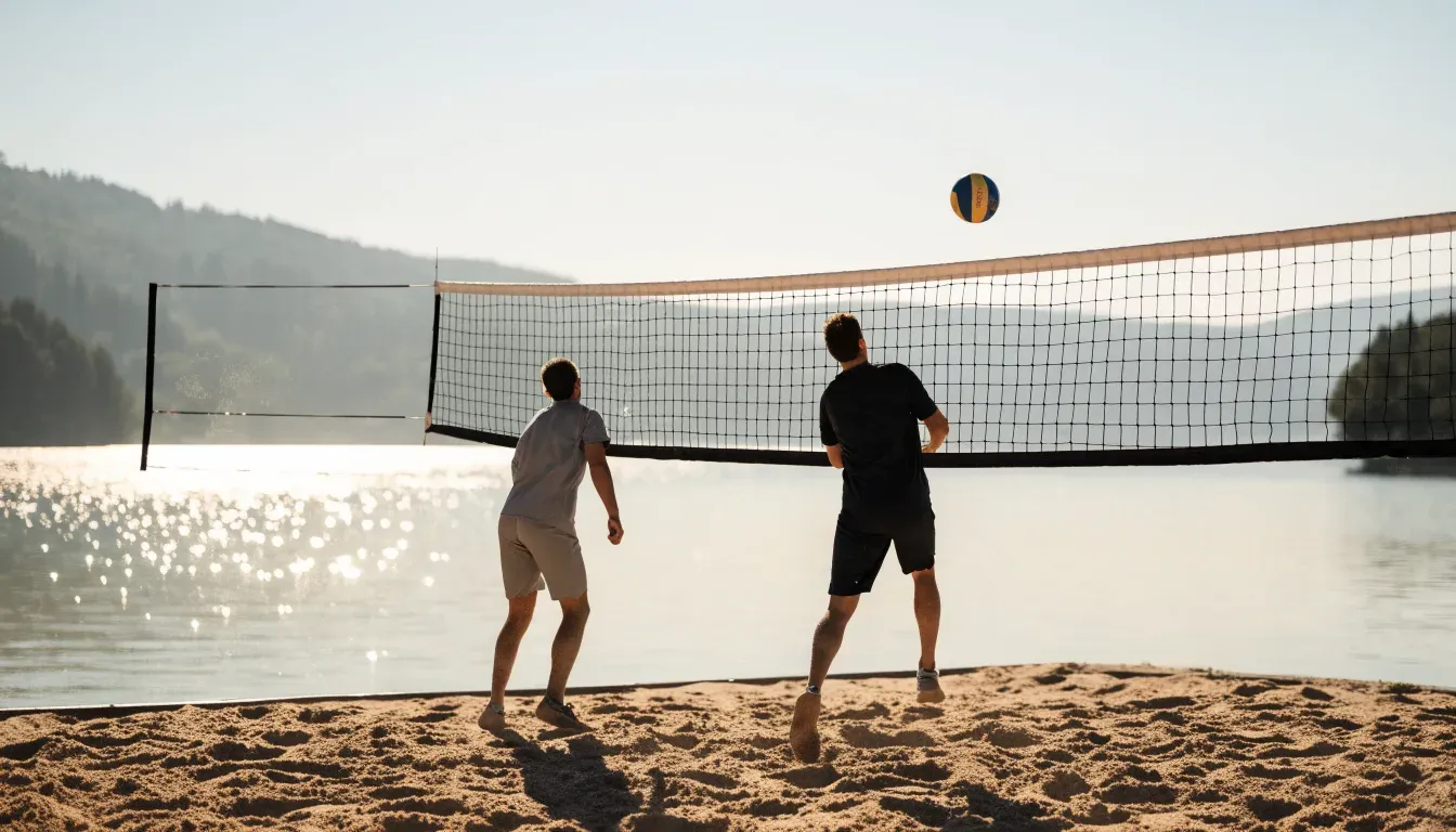 Ein Volleyball liegt auf dem Sand eines Strandfeldes am Seeufer, im Hintergrund glitzert das Wasser.