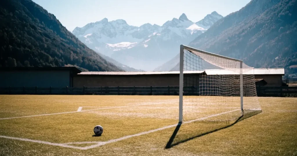 Ein Dorf-Sportplatz mit einem Ball am Strafraumrand, dahinter schneebedeckte Alpengipfel im klaren Morgenlicht