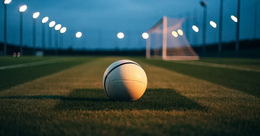 Ein Fussball liegt auf dem Anstosspunkt eines leeren Rasenplatzes in der Abenddämmerung, im Hintergrund leuchten Flutlichtmasten.