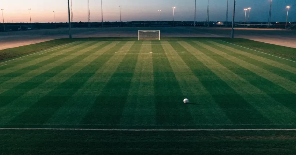 Ein menschenleerer Fussballplatz mit frisch gemähtem Rasen unter Flutlichtmasten in der Abenddämmerung, aus erhöhter Perspektive fotografiert.